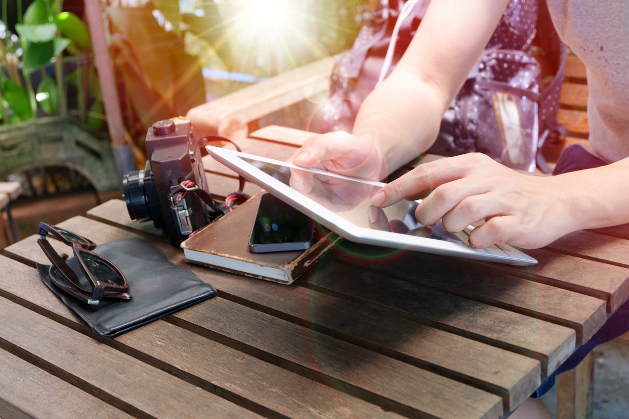 the-story Person using a tablet outdoors alongside a camera, phone, sunglasses, and notebook on a wooden table.